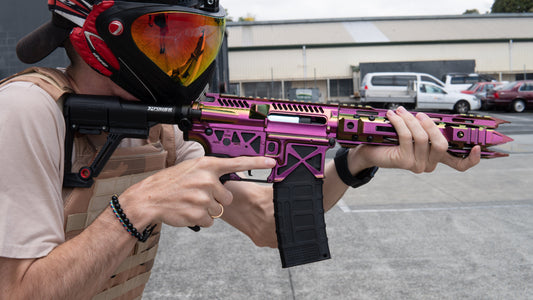 Person in tactical gear aiming a custom purple and gold gel blaster outdoors, wearing a red and black protective mask and tan plate carrier vest in a parking lot setting.